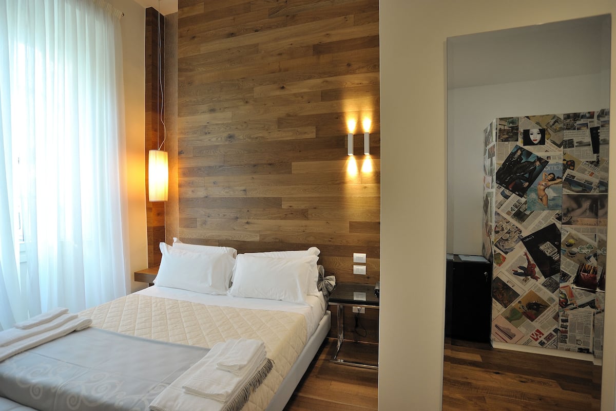 Een hotelkamer met een houten hoofdbord, een bed met witte lakens, een nachtkastje met een lamp, en een muur met een foto-collage.
