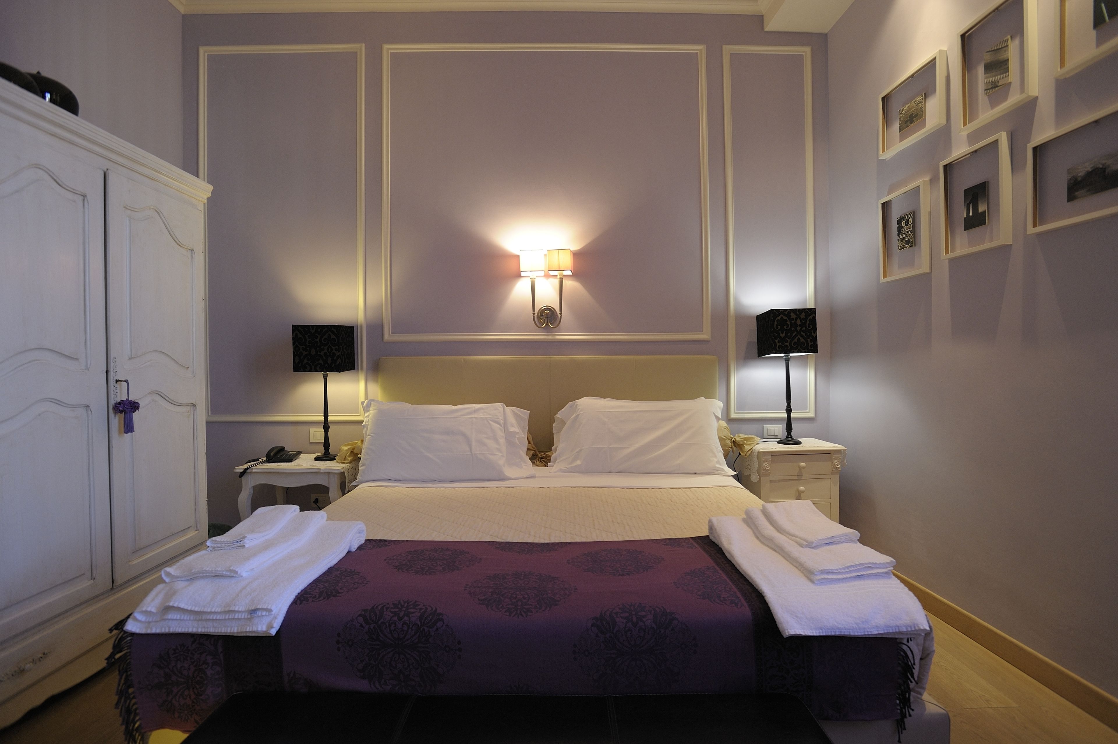 A neatly made bed with white linens, two bedside tables with lamps, a wardrobe, and framed pictures on the wall.
