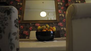 A room with a floral wallpaper, a framed mirror, a bowl of fruit on a table, and a chair.