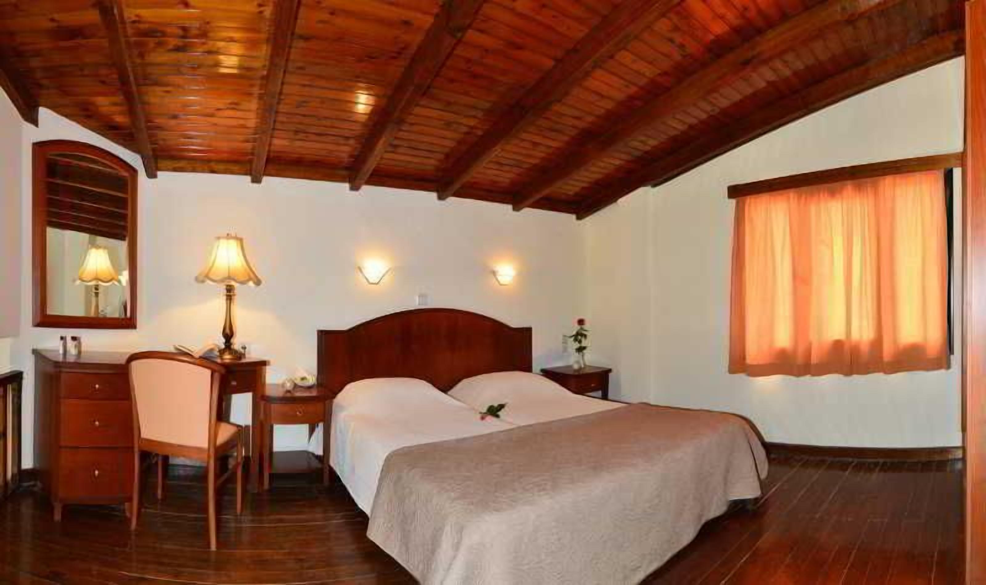 Habitación de hotel con techo de madera, cama con cubrecama color beige, escritorio con lámpara, una silla y una mesita de noche.