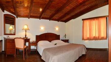 Habitación de hotel con techo de madera, cama con cubrecama color beige, escritorio con lámpara, una silla y una mesita de noche.