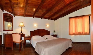 A hotel room with a wooden ceiling, a bed with a beige bedspread, a desk with a lamp, a chair, and a nightstand. - Antilia Apts (Platanias)