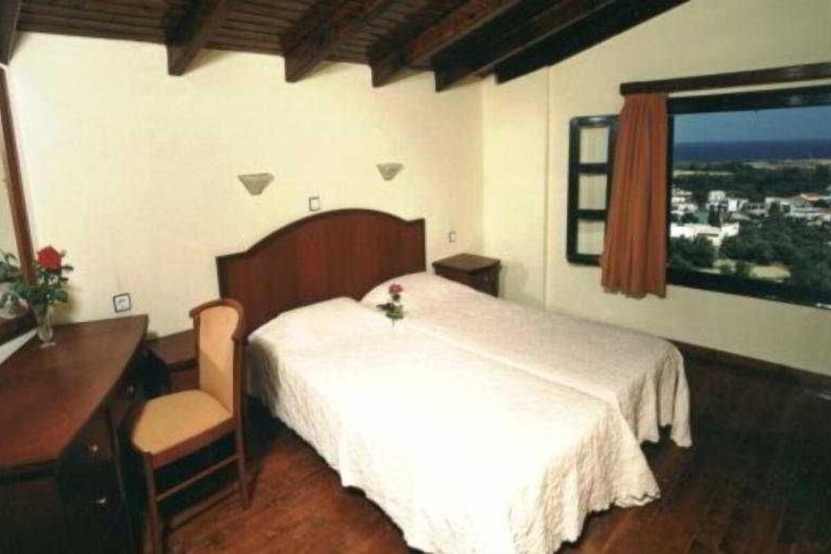 A bedroom with a wooden bed, a desk, and a chair. There is a window with a view of buildings and a curtain.