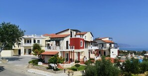 A multi-story building with a red and white facade, a visible swimming pool, and a clear view of the sea in the background. - Antilia Apts (Platanias)