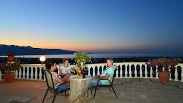 A terrace with a table set for two, overlooking a body of water and mountains at dusk.