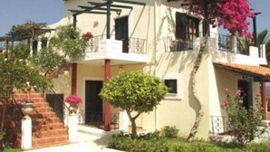 A two-story building with a balcony, a staircase, and a garden with flowering plants. - Antilia Apts (Platanias)