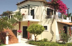 A two-story building with a balcony, a staircase, and a tree in the front yard. - Antilia Apts (Platanias)