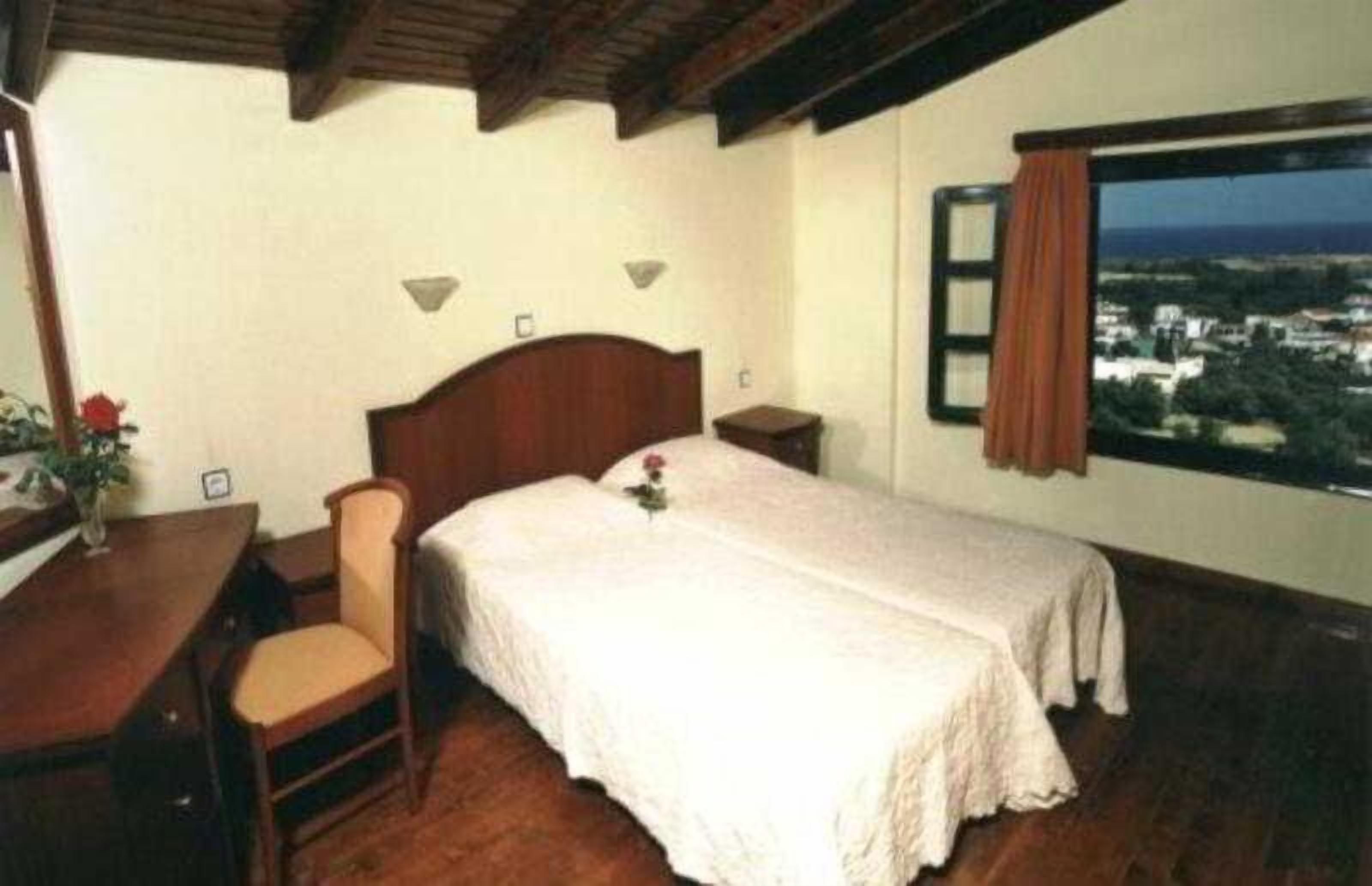 A bedroom with a wooden bed, a desk with a chair, a window with a view, and a nightstand.