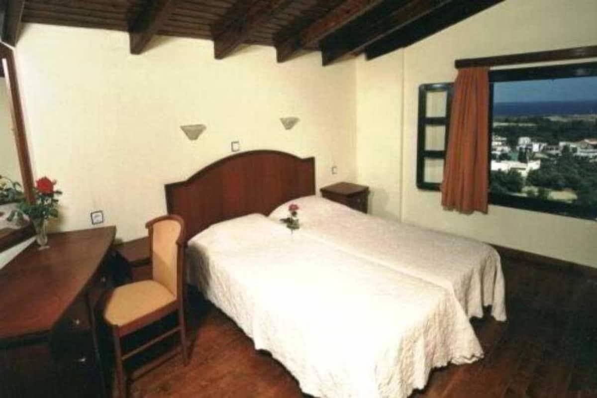 A bedroom with a wooden bed, a desk, and a chair. There is a window with a view of buildings and a curtain.
