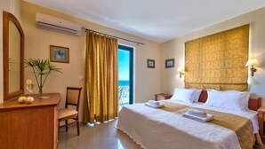 A hotel room with a bed, a desk with a chair, a potted plant, and a view of the sea through a window. - Antilia Apts (Platanias)