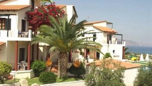 A two-story building with balconies, a palm tree, and a view of the sea. - Antilia Apts (Platanias)