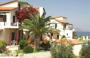 A two-story building with a balcony, a palm tree, and a view of the sea. - Antilia Apts (Platanias)
