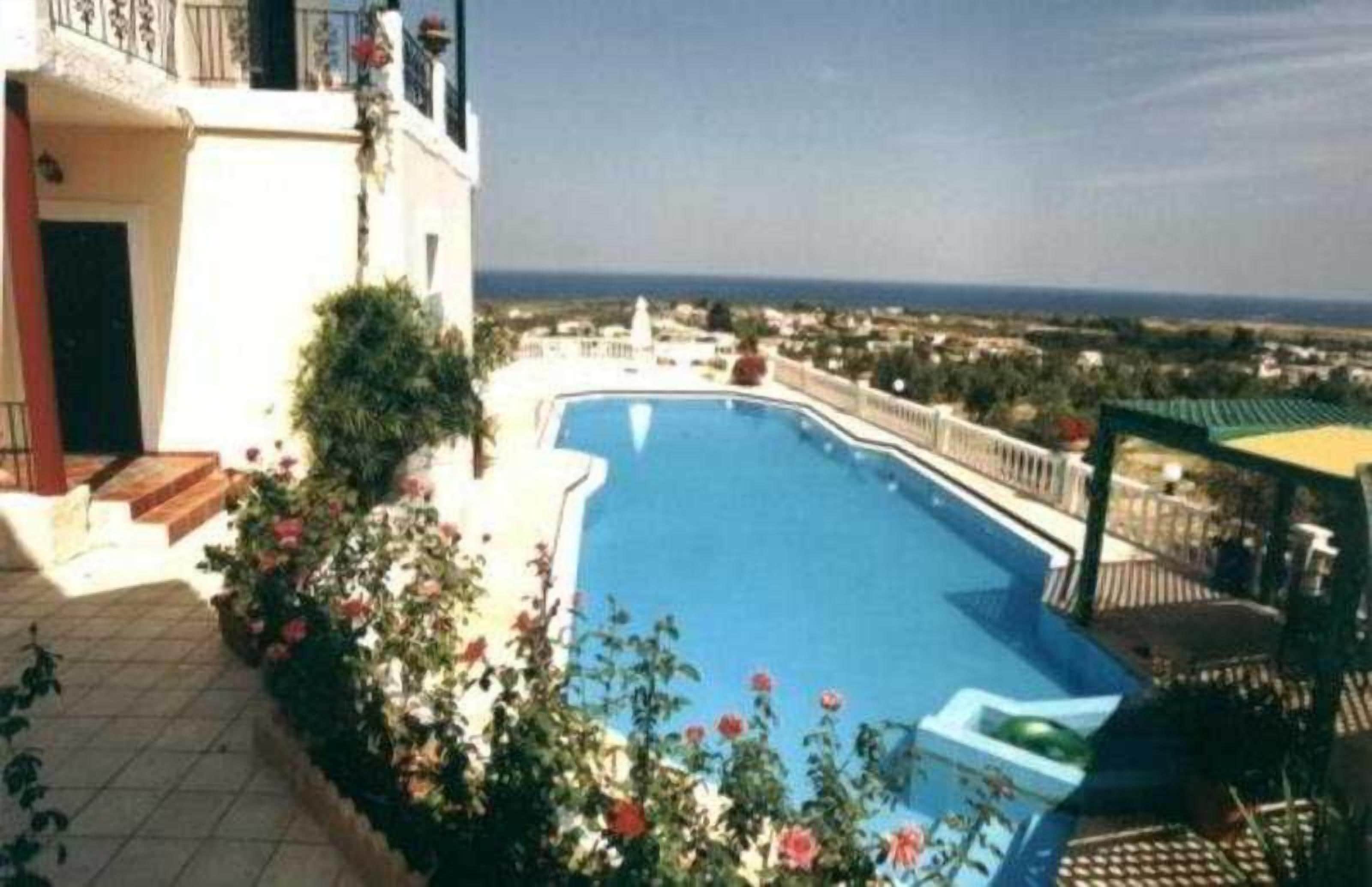 A swimming pool with a view of the sea, surrounded by a tiled patio and some greenery.