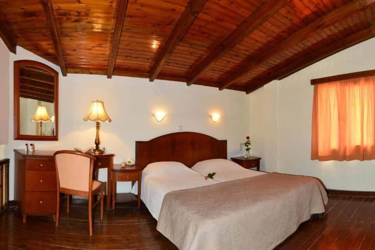 A bedroom with a wooden ceiling, a bed with a beige bedspread, a desk with a lamp, a chair, and a nightstand with a vase.