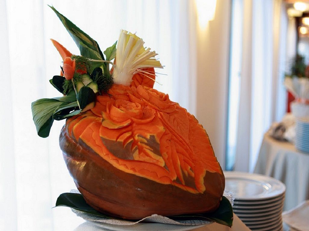 A carved pumpkin with a decorative top, placed on a white surface with a blurred background of a room with tables and chairs.