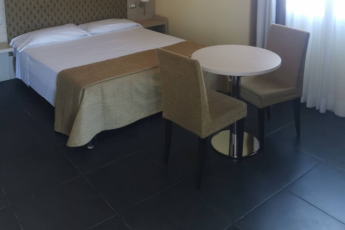 Habitación de hotel con cama, una mesa redonda, dos sillas, una mesita de noche, una lámpara y dos cuadros colgados en la pared.