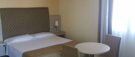 A hotel room with a bed, a round table, two chairs, a nightstand, a lamp, and two wall-mounted pictures.