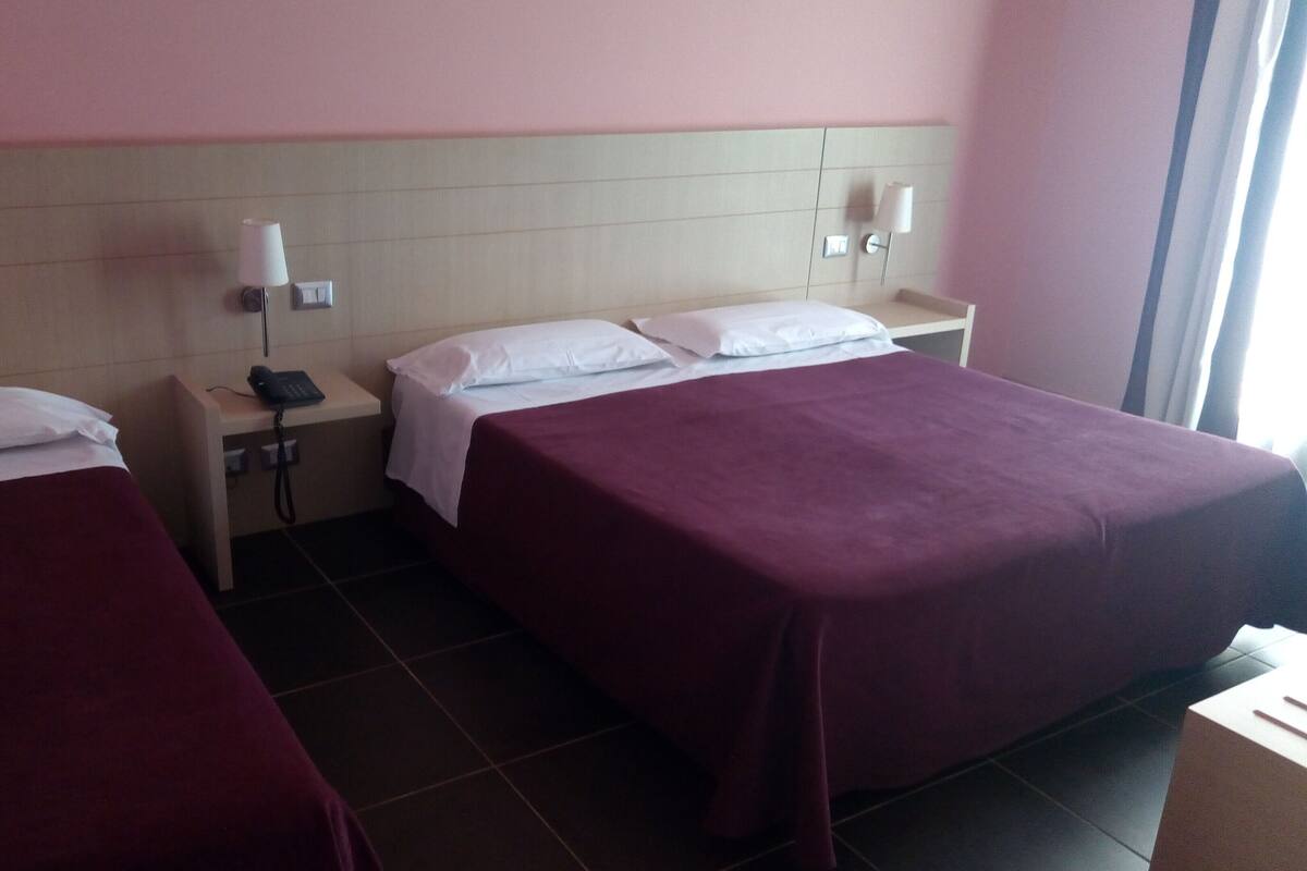 Habitación de hotel con dos camas, cada una con un almohadón blanco y una colcha morada. Hay dos mesitas de noche, cada una con una lámpara y un teléfono. El piso es de baldosas y la pared es rosa.
