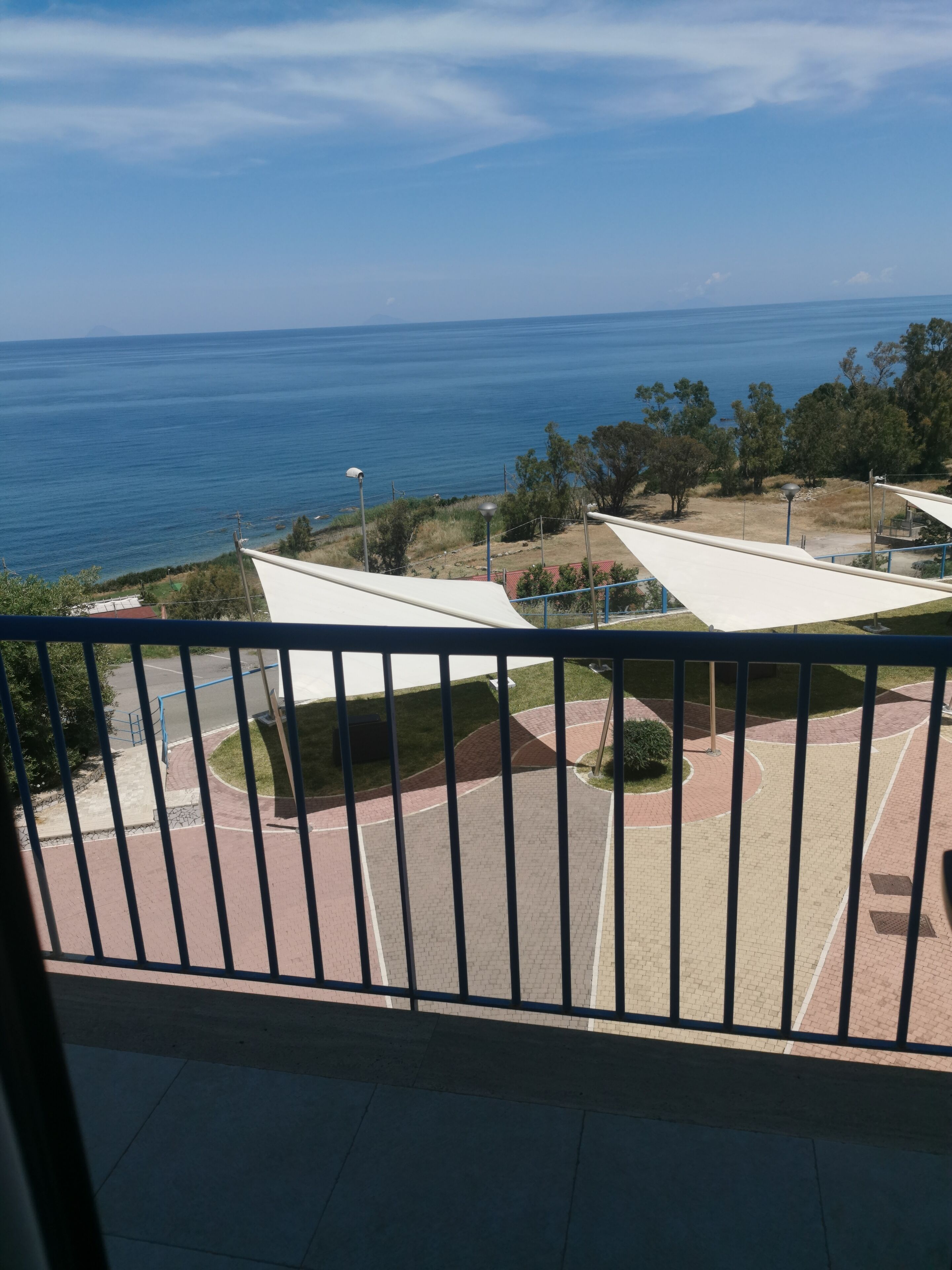 Un balcone con vista sul mare, una zona pavimentata con un lampione e una distesa erbosa alberata.