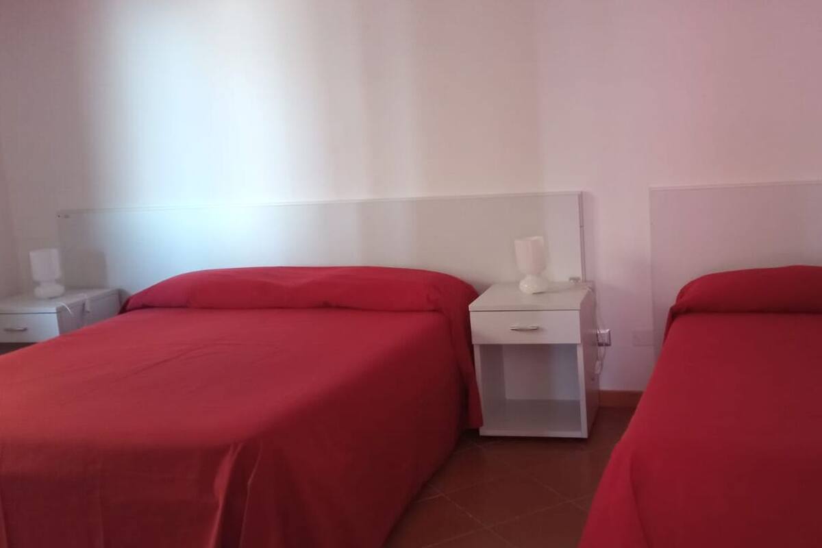Una habitación con dos camas individuales, cada una con una colcha roja y cabeceros blancos. Entre las camas hay una mesita de noche con una lámpara.