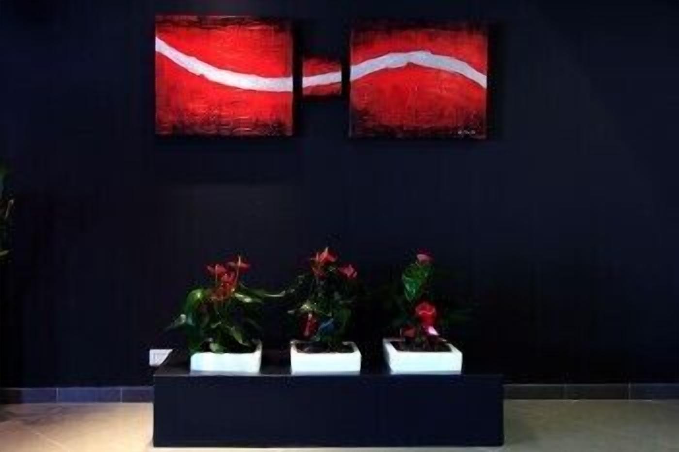 A reception area with a dark wall, two red and white abstract paintings, and three potted plants with red flowers.