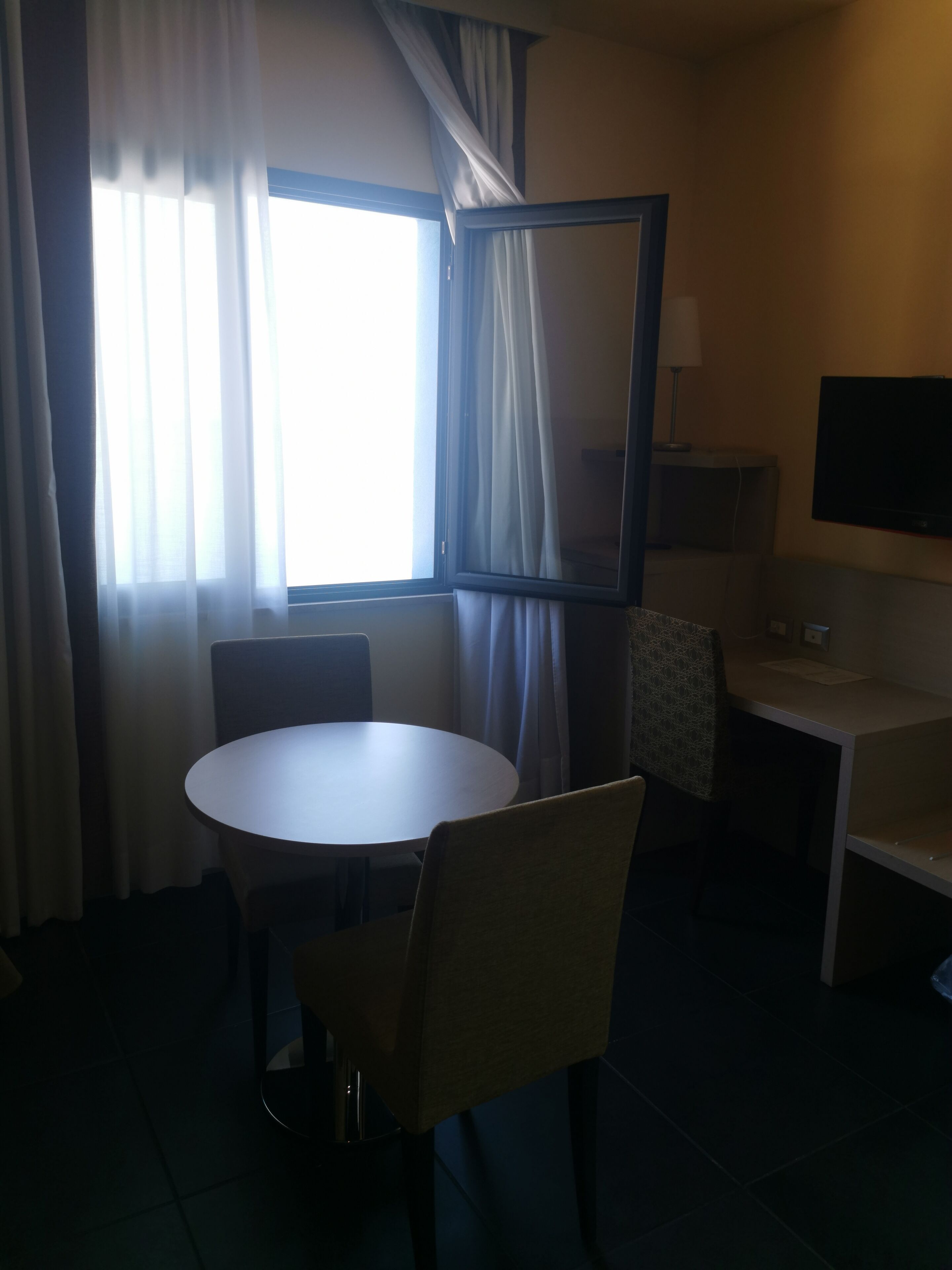 A hotel room with a round table, two chairs, a TV on a stand, and a window with curtains.
