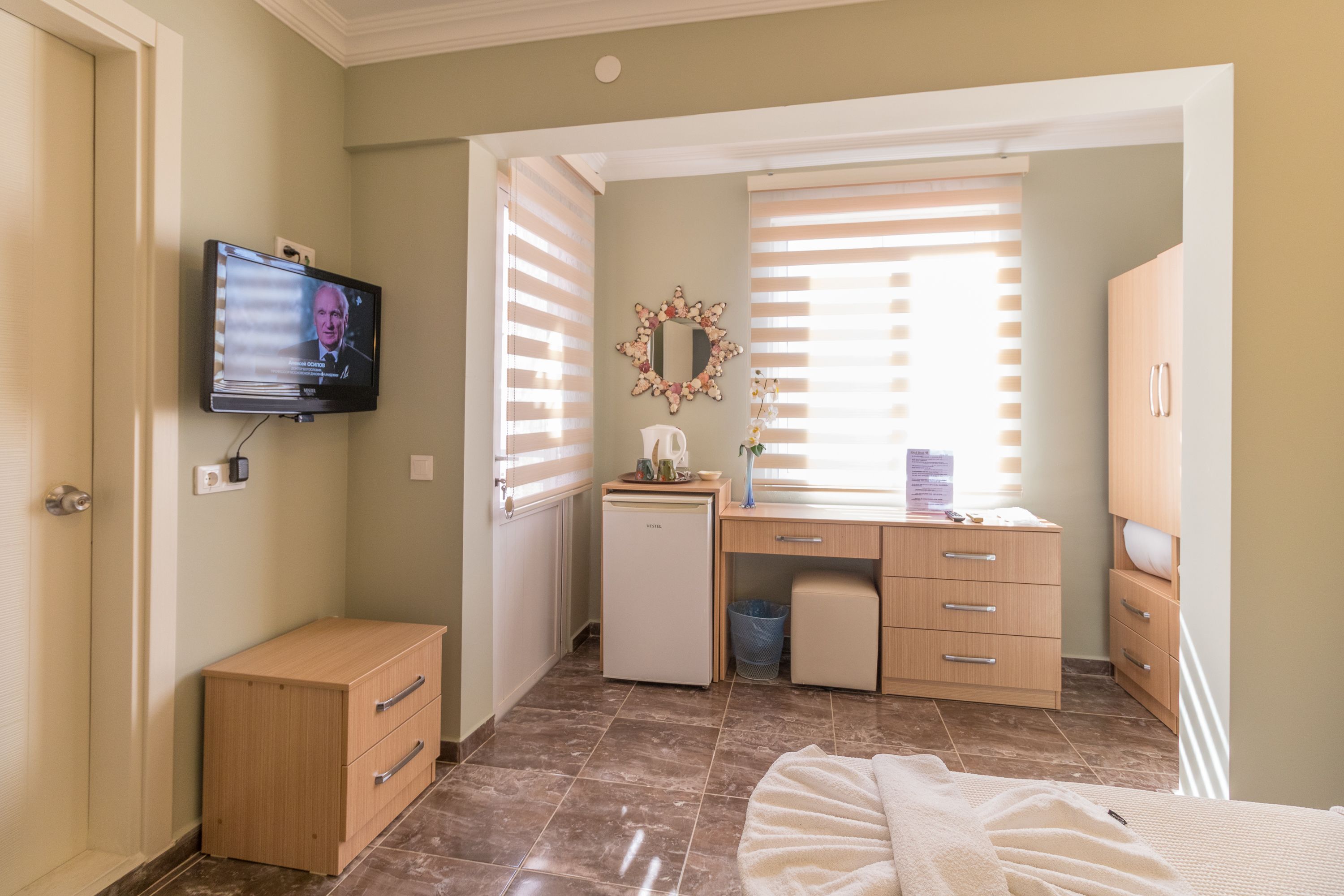 A bedroom with a bed, a dresser, a desk with a computer, a small refrigerator, and a TV mounted on the wall.