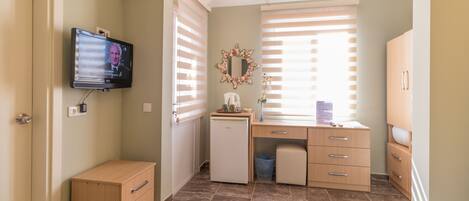 A bedroom with a bed, a dresser, a desk with a computer, a small refrigerator, and a TV mounted on the wall.