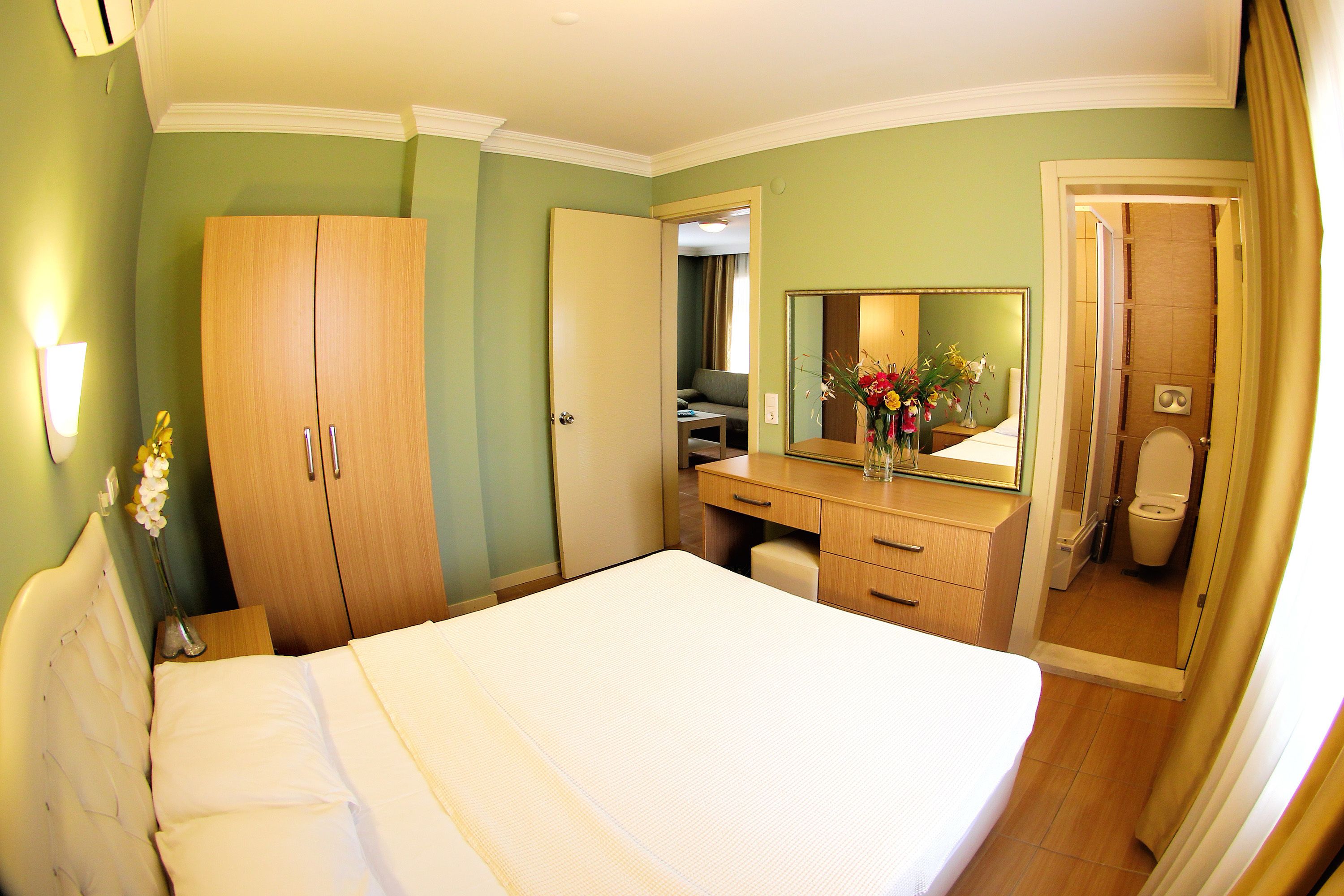 A hotel room with a bed, a wooden wardrobe, a desk with a mirror, and a bathroom visible through an open door.