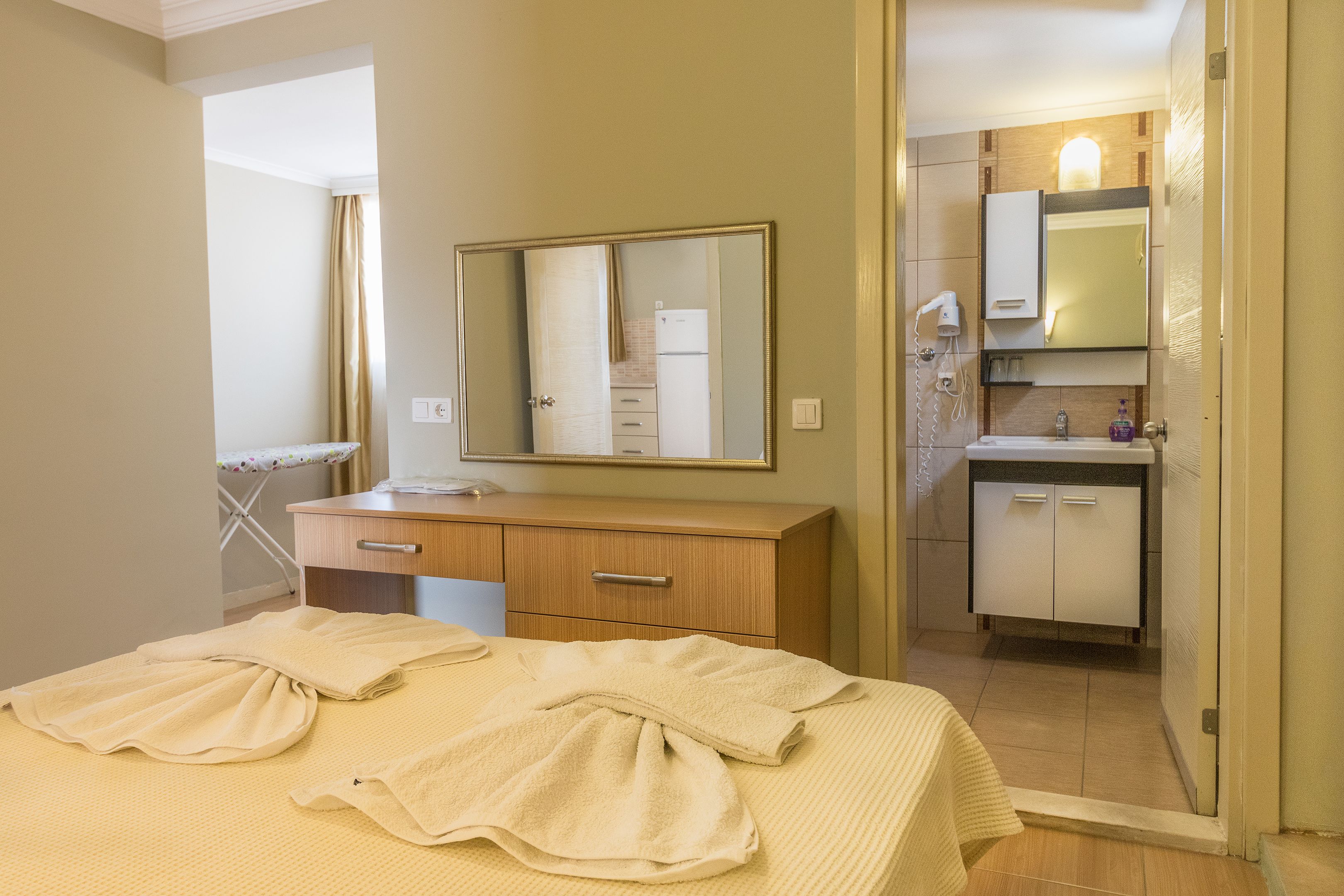 Habitación de hotel con cama, cómoda de madera, espejo, baño con lavamanos y espejo, y ventana con cortinas.