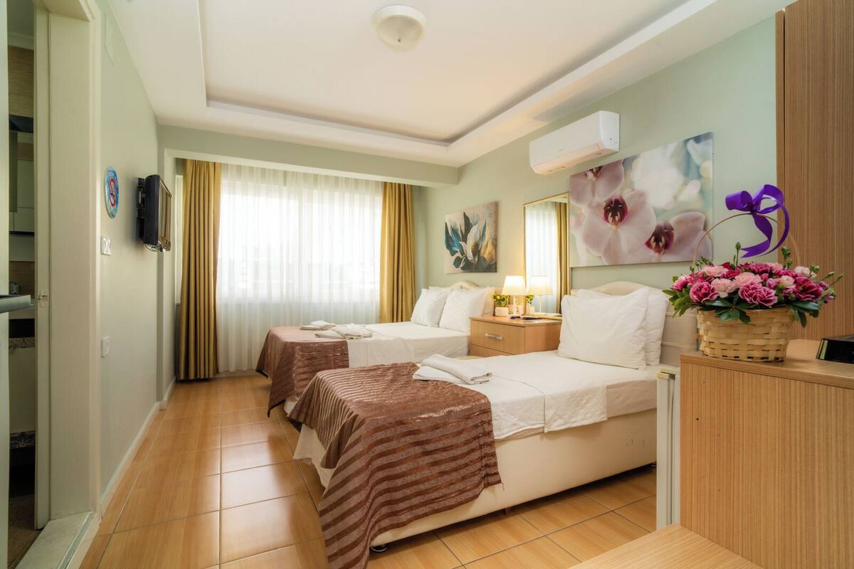 A hotel room with two beds, a bedside table with a lamp, a wall-mounted air conditioner, and a floral decoration.