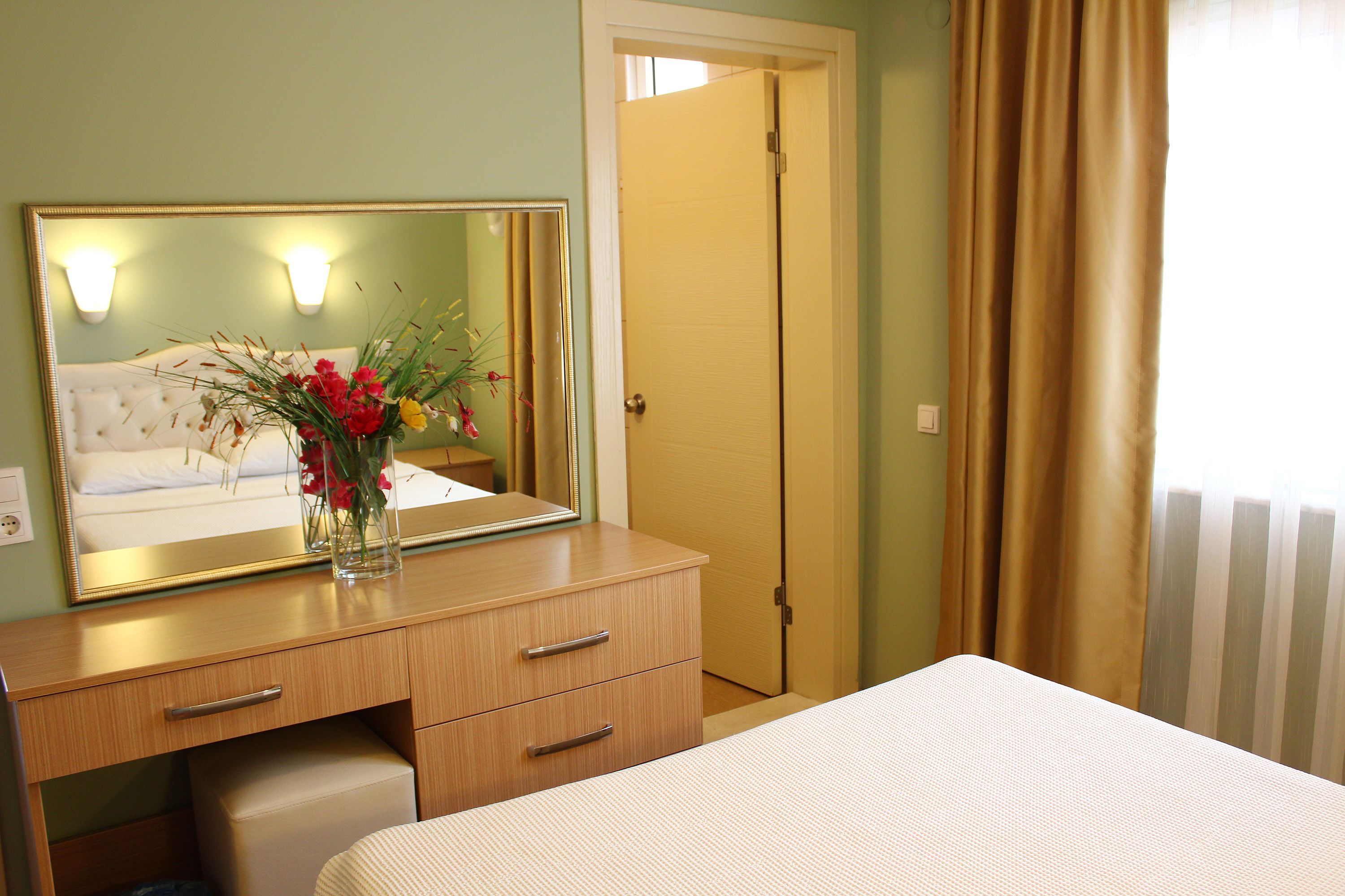 A hotel room with a bed, a wooden dresser with drawers, a mirror, and a vase with flowers.