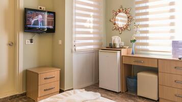 A hotel room with a television mounted on the wall, a desk with a computer, a small refrigerator, a bedside table with a vase of flowers, and a window with blinds.