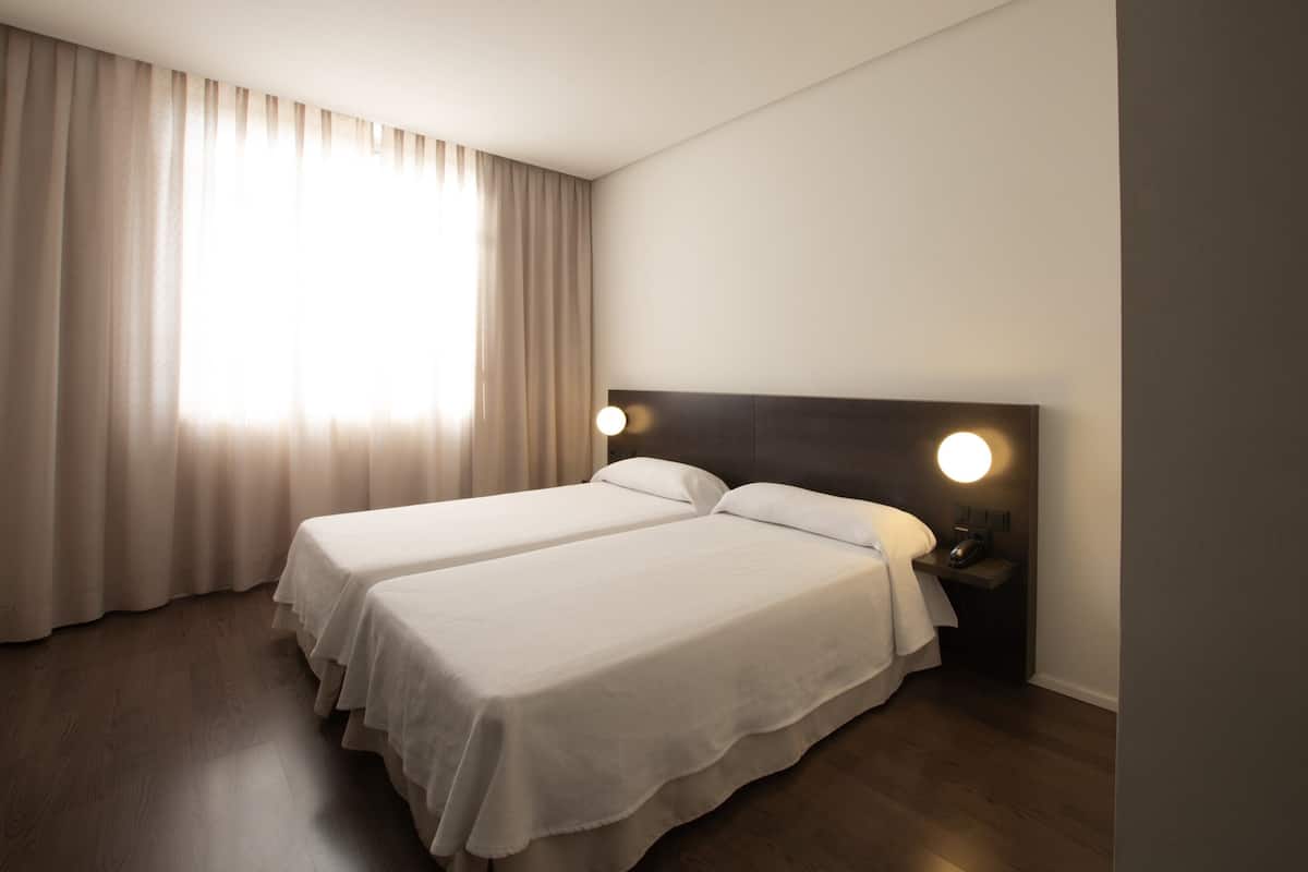 A hotel room with two beds, a large window with curtains, a bedside table with a lamp, and wooden flooring.