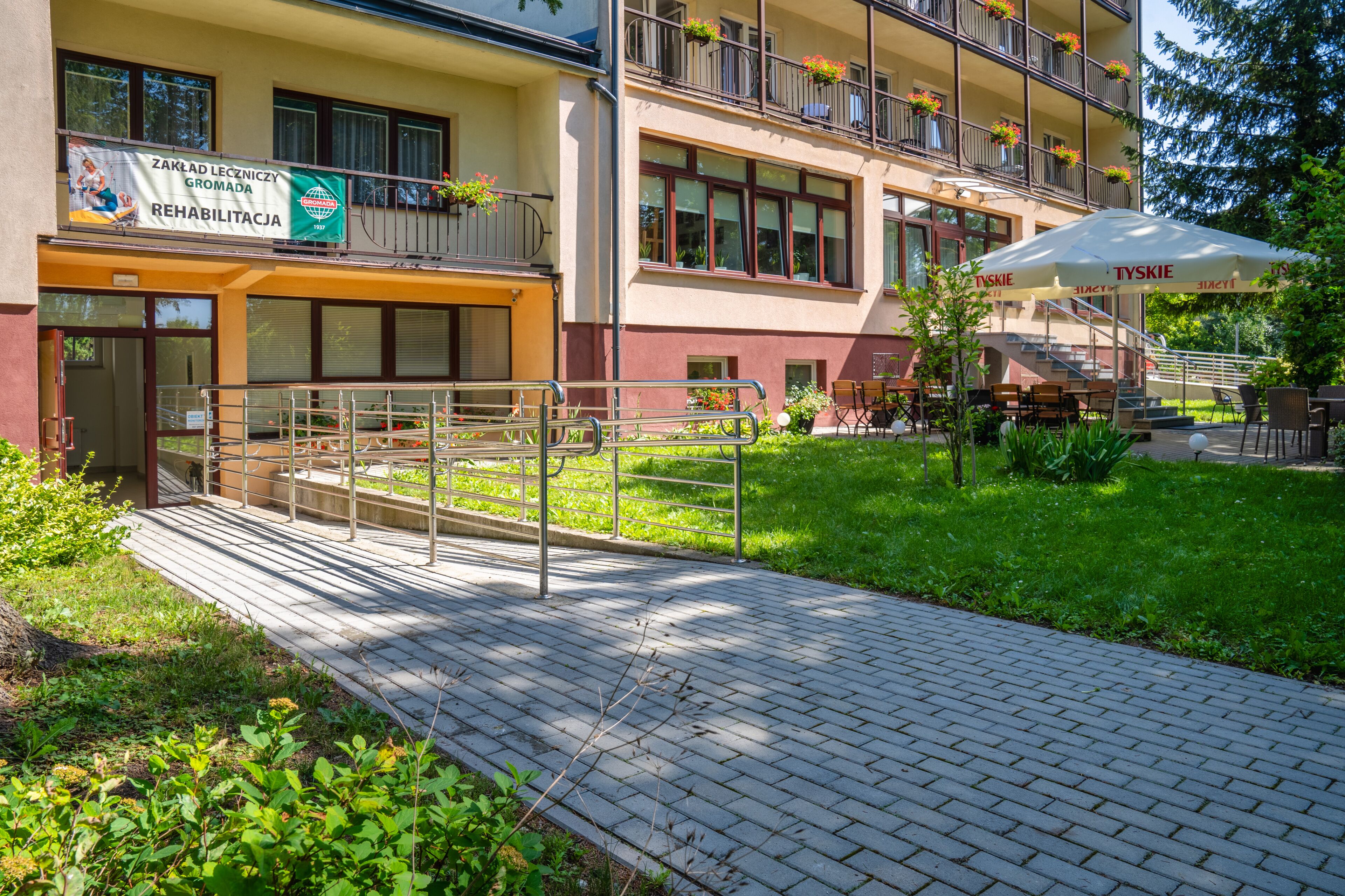 Un edificio con un cartello che riporta 'Zakład Leczniczy Gromada REHABILITACJA' e posti a sedere all'aperto con ombrelloni.
