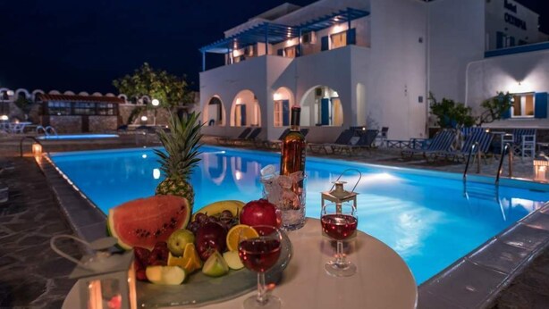 A poolside setting with a table of fruits and drinks, a pool, and a building with arched windows and a blue roof. - Hotel Olympia (Santorini)