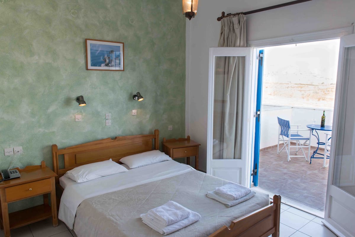 A bedroom with a bed, bedside tables, a nightstand, a window with curtains, and a view of an outdoor area with a table and chairs.