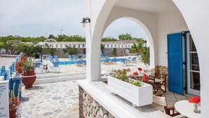 A poolside area with a stone floor, blue and white lounge chairs, and a white planter with red flowers. - Hotel Olympia (Santorini)