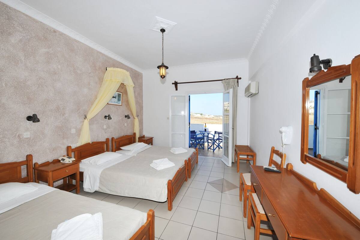 A hotel room with two beds, a wooden dining table, and a balcony with blue chairs.