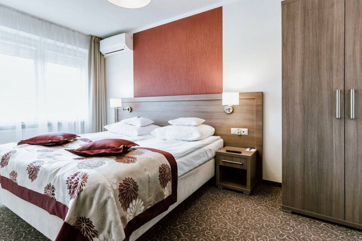 A hotel room with a bed, bedside table, lamp, wardrobe, and a large window with curtains.