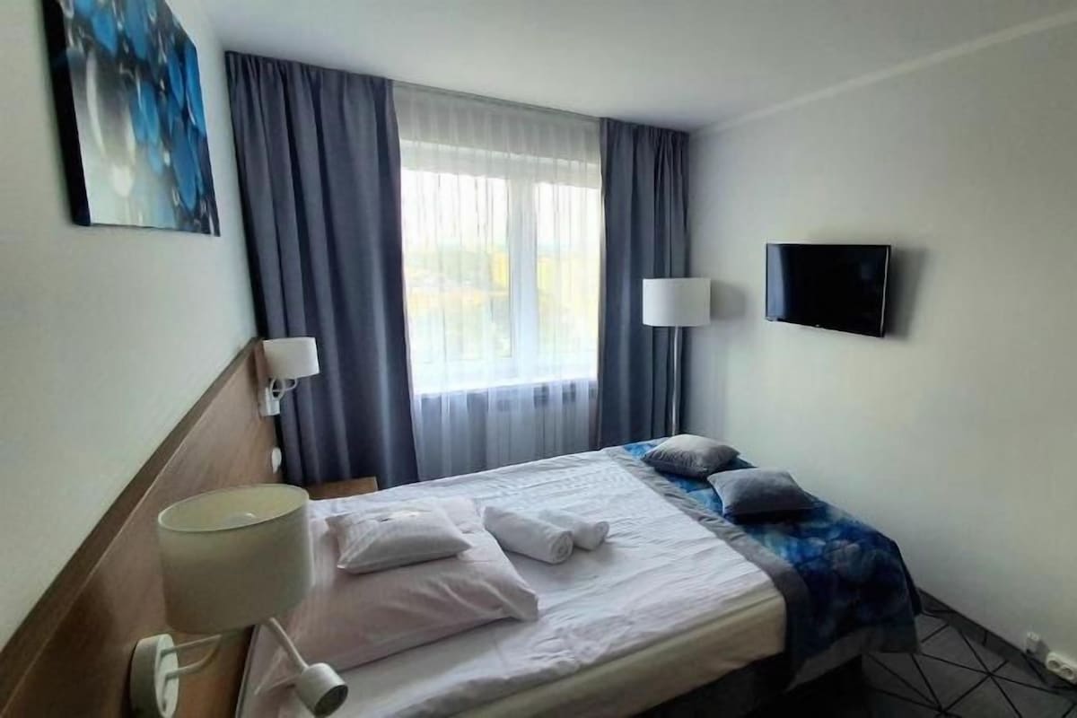 A hotel room with a bed, a television, a lamp, and a window with curtains.