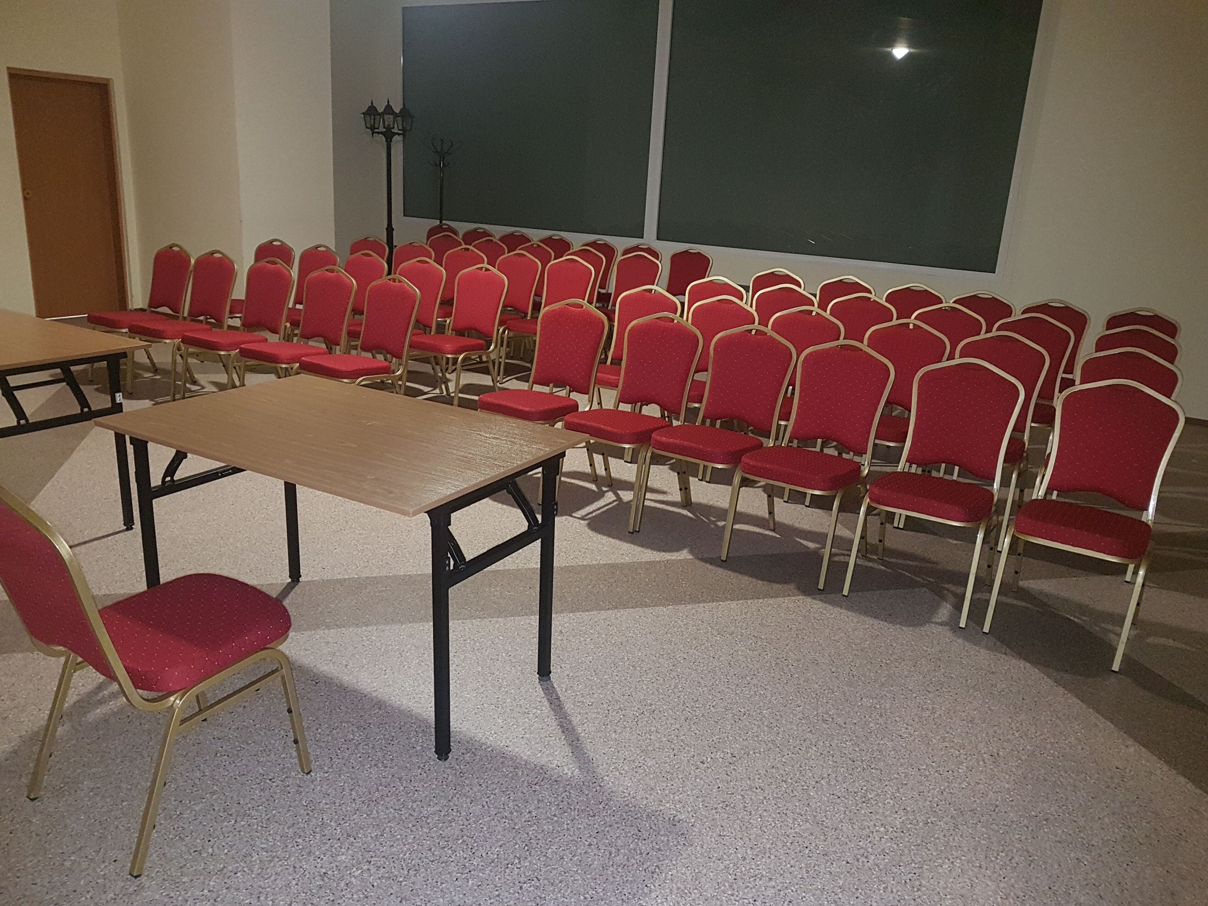 A conference room with rows of red chairs and a long table, a projector screen, and a lamp.