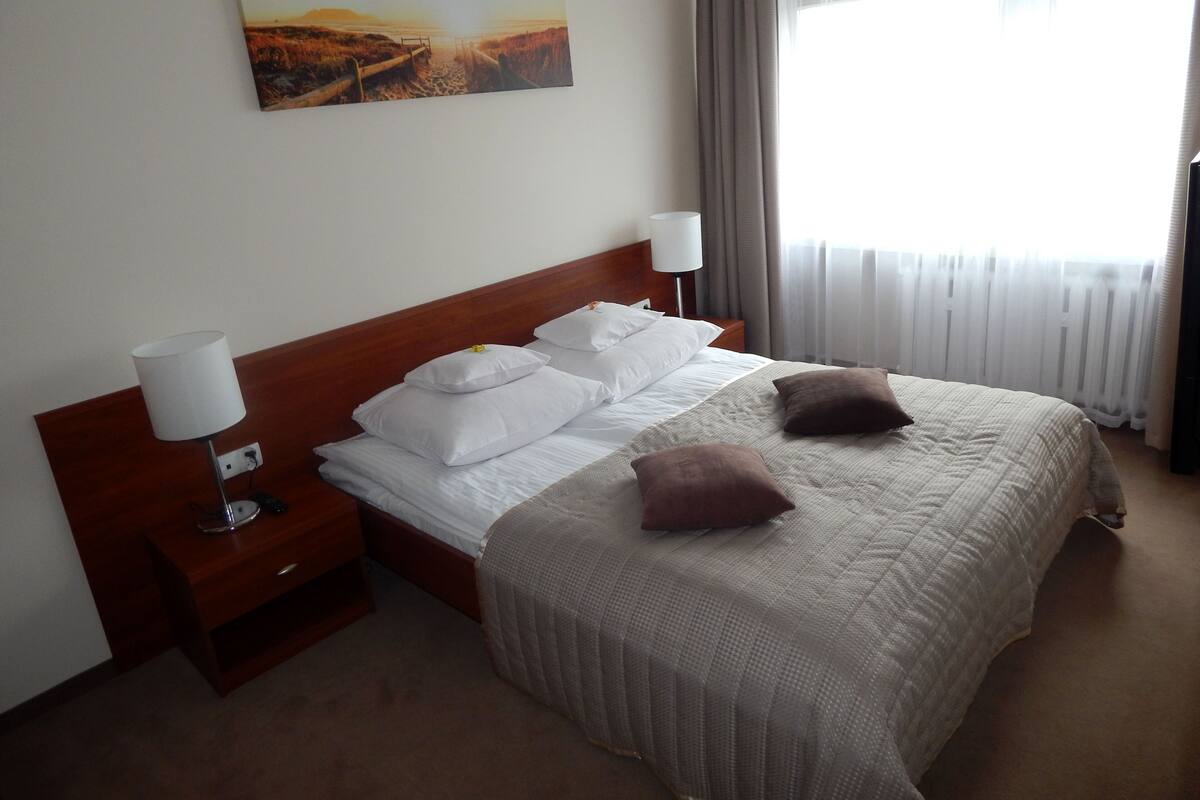 A hotel room with a wooden headboard, a large bed with white and brown pillows, a bedside table with a lamp, and a painting of a sunset over a beach on the wall.