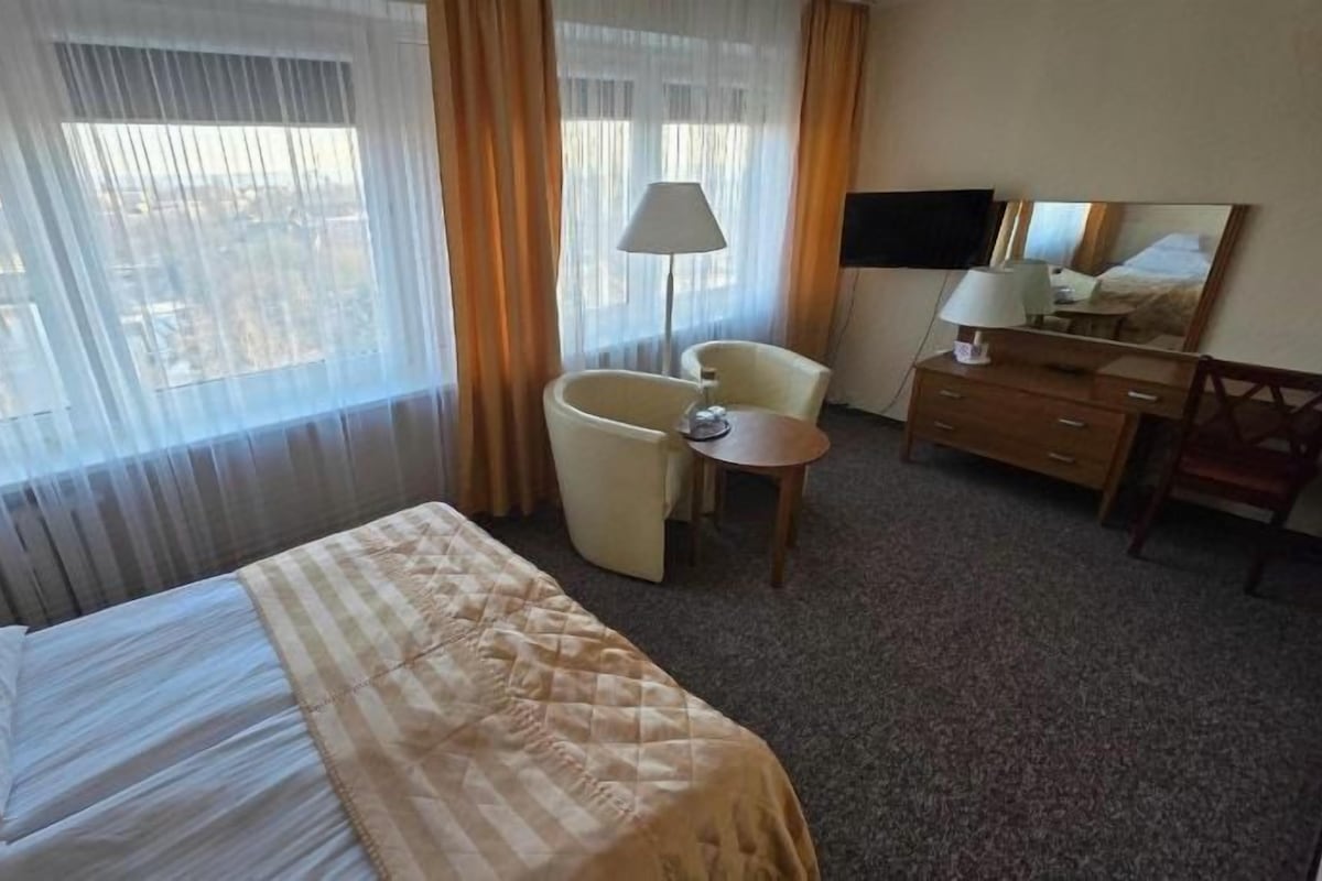 A hotel room with a bed, a chair, a small table, a desk, a lamp, a television, and a window with curtains.