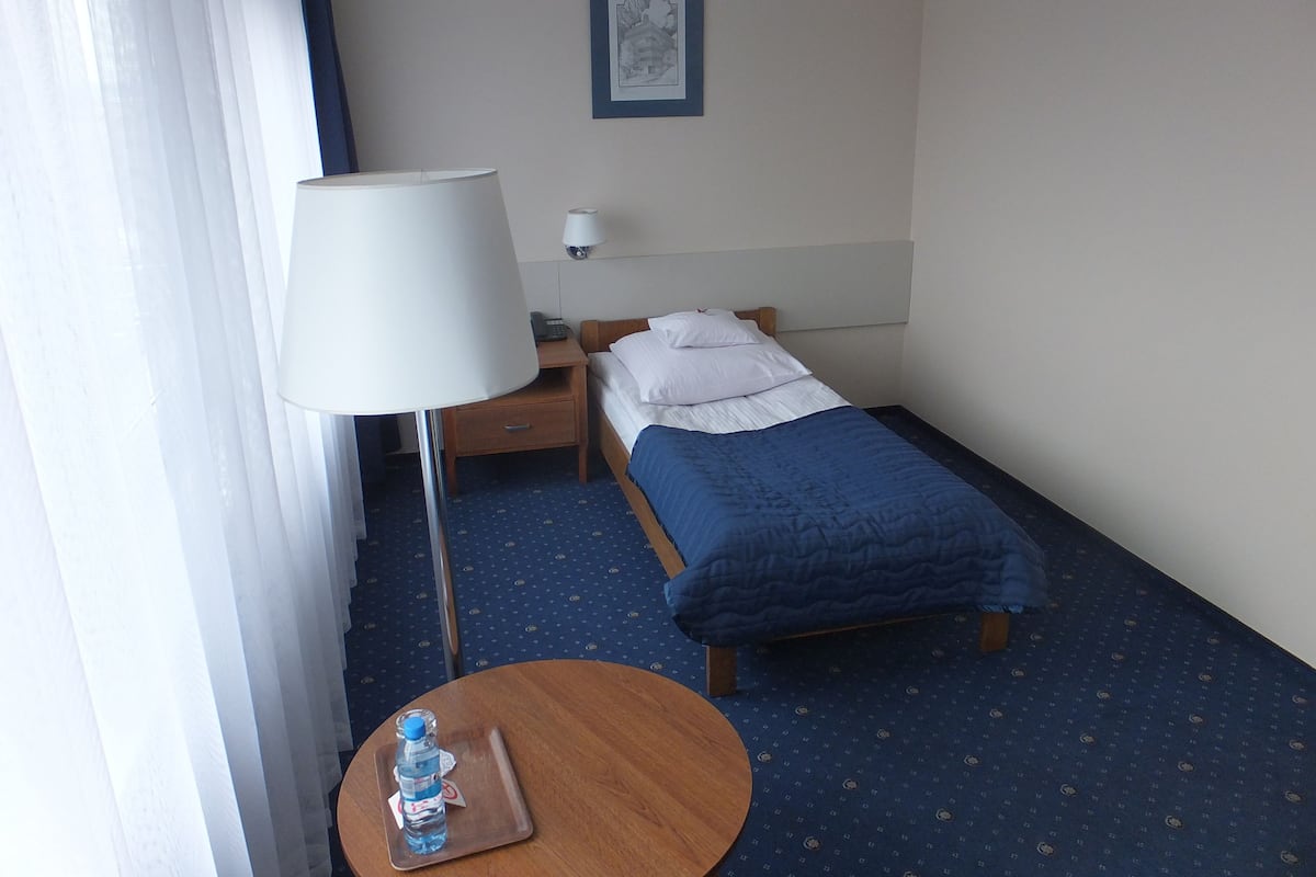 A hotel room with a single bed, a bedside table with a lamp, a small round table with a water bottle, and a framed picture on the wall.