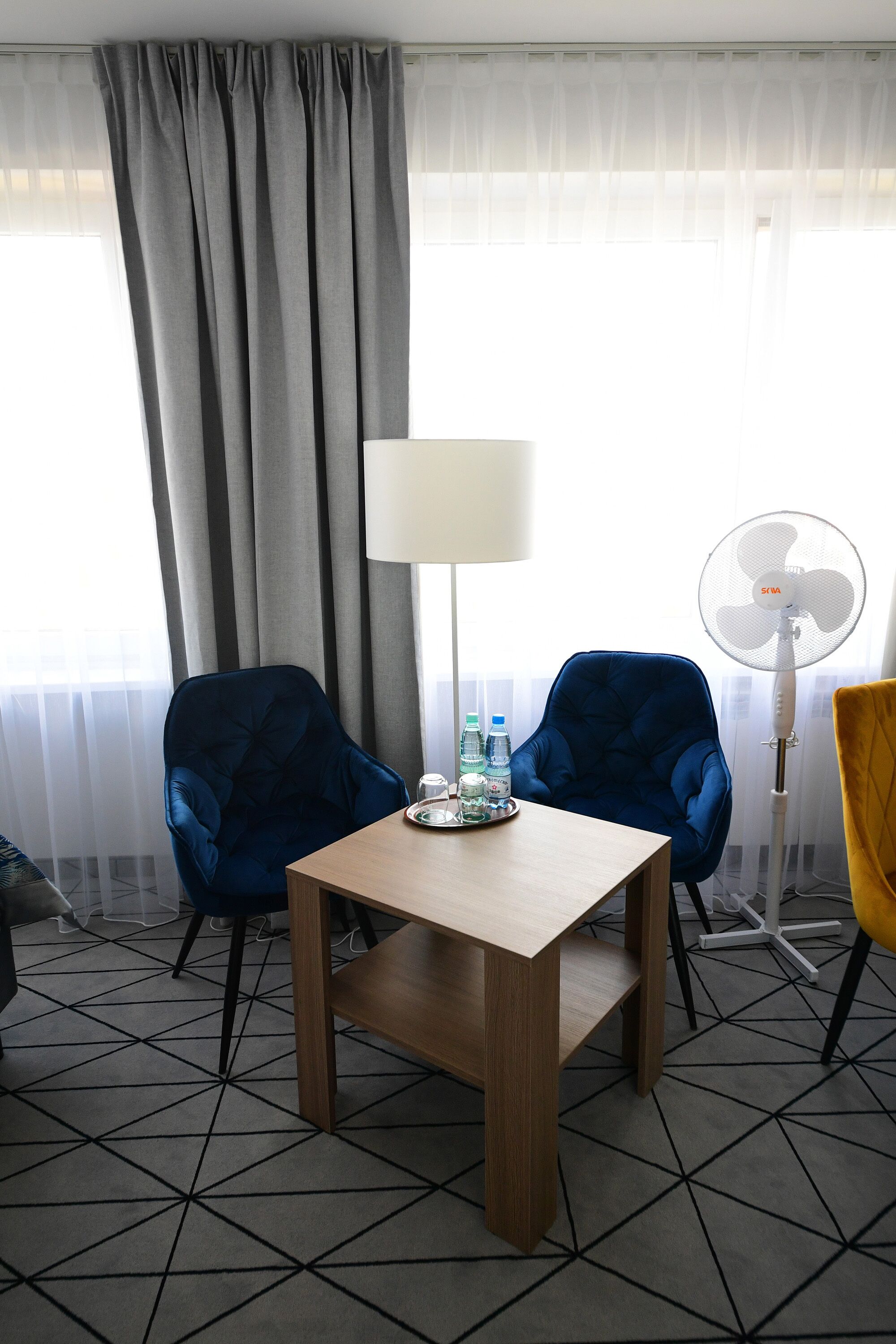 Une chambre d’hôtel avec une table en bois, deux fauteuils bleus, une lampe, un ventilateur et une bouteille d’eau posée sur la table.
