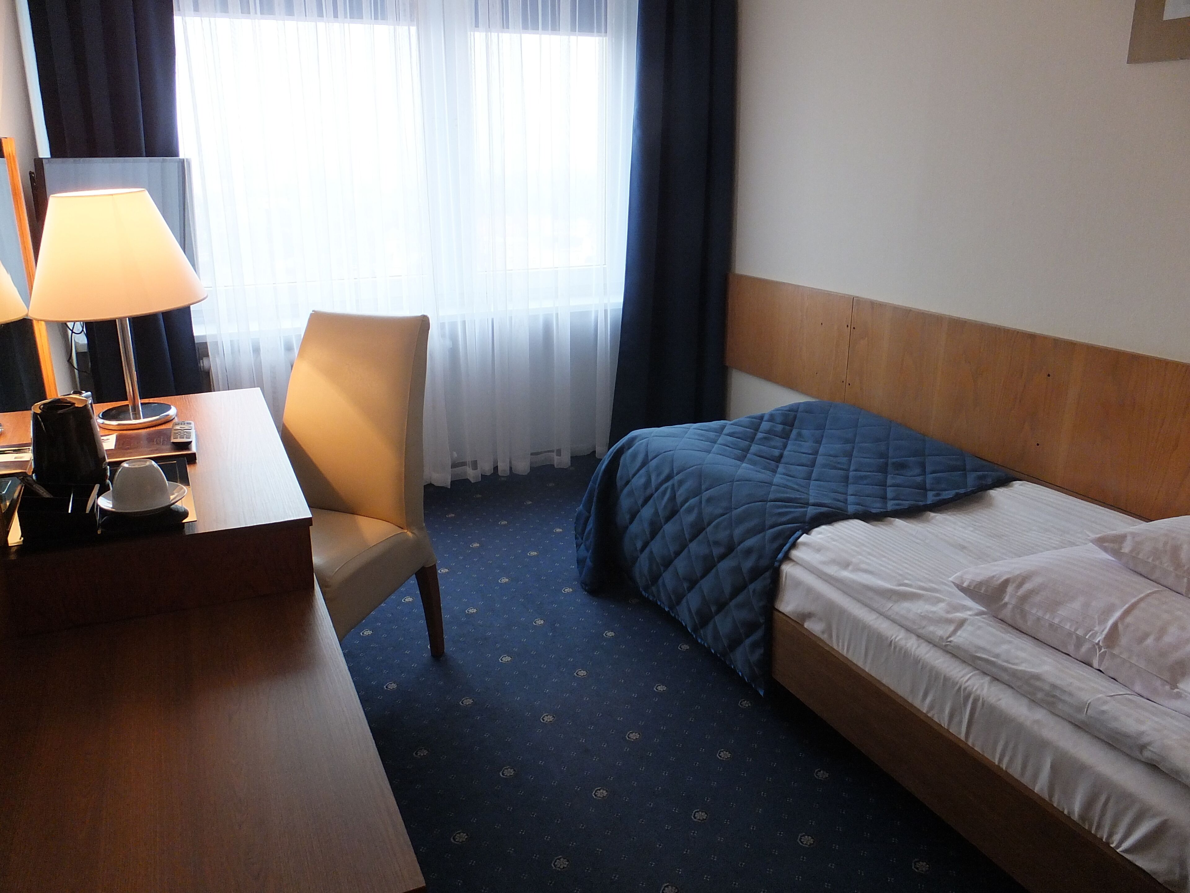 Une chambre d’hôtel avec un lit, un bureau doté d’une lampe, une chaise et une fenêtre avec des rideaux.