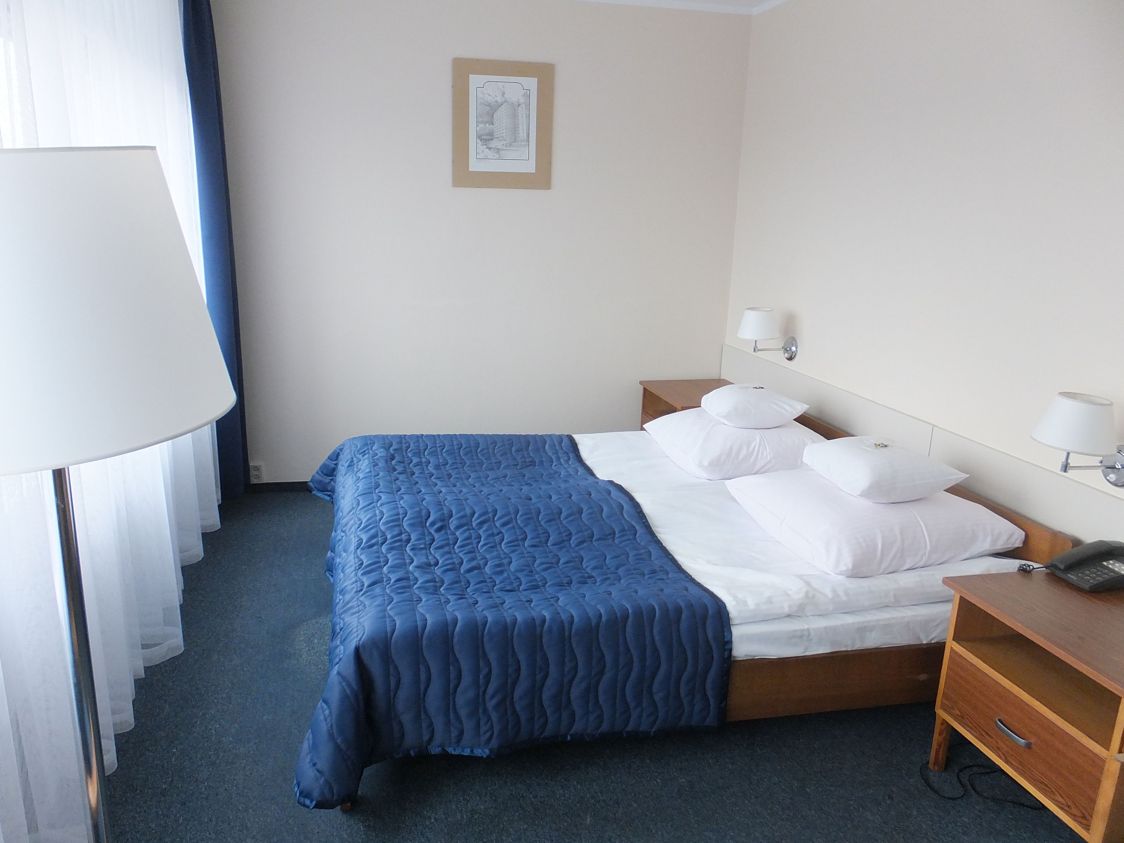 Habitación de hotel con cama, lámparas de noche, mesita de noche y un cuadro en la pared.