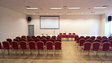 Une salle de conférence avec des rangées de chaises rouges, un grand écran et une table équipée d’un microphone.