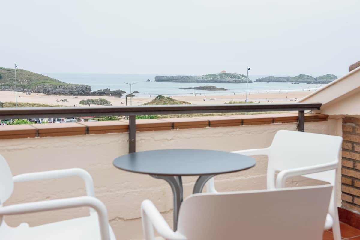 A balcony with a table and chairs overlooking a beach with rocky outcrops and a distant view of the sea.
